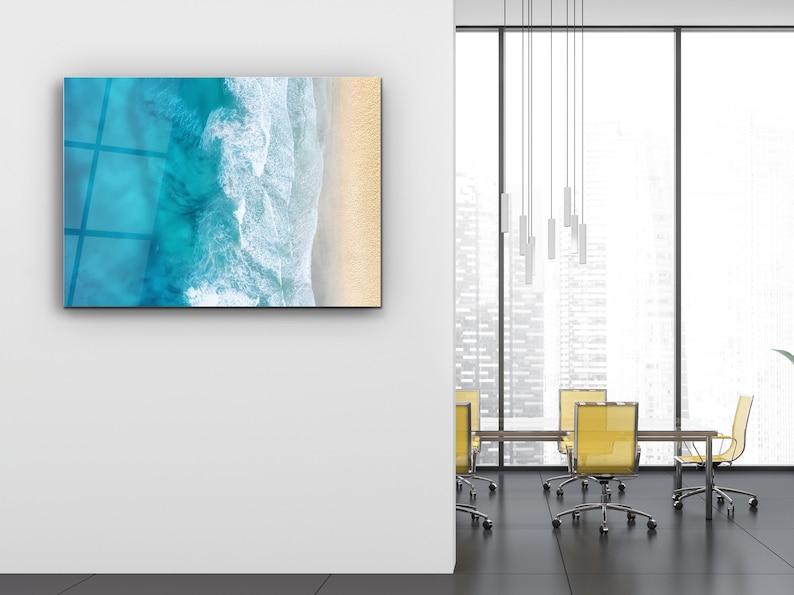 May include: A modern office space with a large window overlooking a city skyline. The room features a glass wall with a framed photograph of an aerial view of ocean waves crashing on a sandy beach. The room is furnished with a wooden table and yellow chairs.