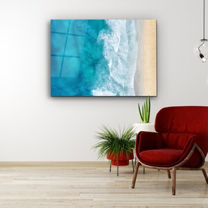 May include: A framed abstract art print of a turquoise ocean with white waves crashing on a sandy beach. The print is hanging on a white wall above a red armchair and a potted plant.