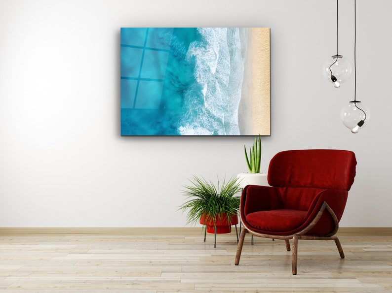 May include: A framed abstract art print of a turquoise ocean with white waves crashing on a sandy beach. The print is hanging on a white wall above a red armchair and a potted plant.