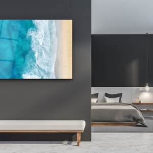 May include: A modern bedroom with a gray and white color scheme. A large framed print of an aerial view of ocean waves crashing on a sandy beach hangs on the wall. A white bench sits in front of the print. A gray bed with white pillows and a gray blanket is in the background.
