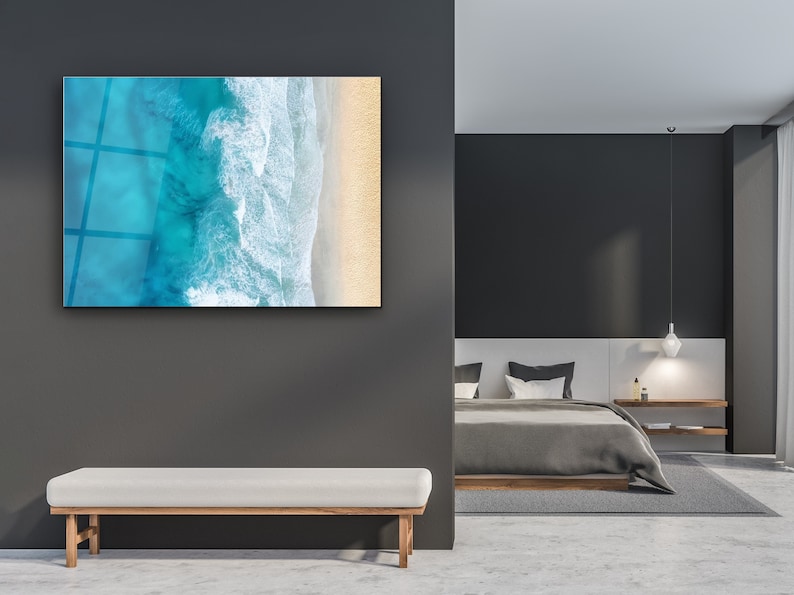 May include: A modern bedroom with a gray and white color scheme. A large framed print of an aerial view of ocean waves crashing on a sandy beach hangs on the wall. A white bench sits in front of the print. A gray bed with white pillows and a gray blanket is in the background.