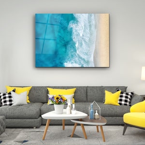May include: A gray sectional sofa with yellow throw pillows in a living room setting. A white coffee table with a vase of yellow flowers sits in front of the sofa. A yellow armchair with a black and white patterned fabric sits in the corner of the room. A framed print of an aerial view of ocean waves crashing on a sandy beach hangs on the wall.