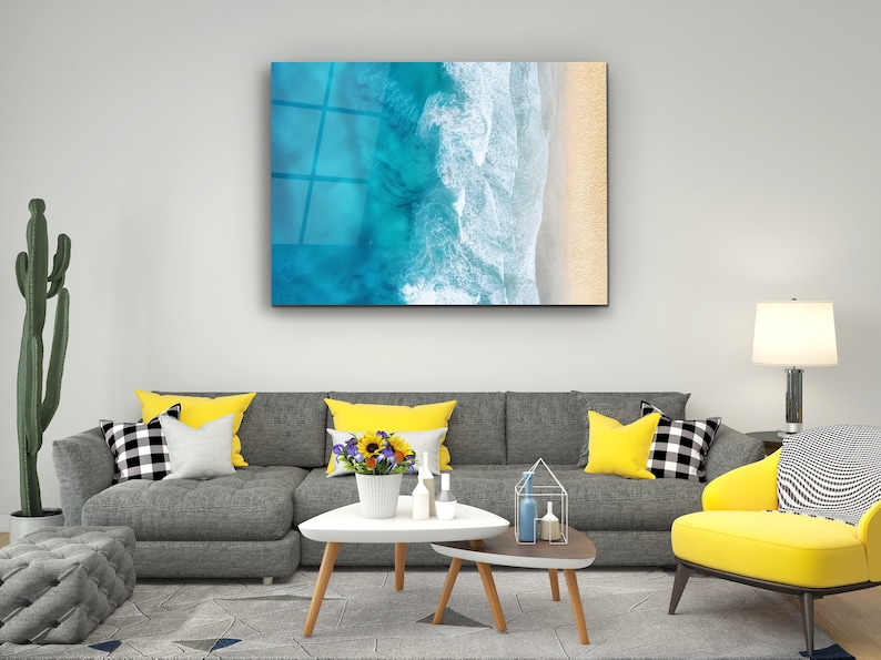 May include: A gray sectional sofa with yellow throw pillows in a living room setting. A white coffee table with a vase of yellow flowers sits in front of the sofa. A yellow armchair with a black and white patterned fabric sits in the corner of the room. A framed print of an aerial view of ocean waves crashing on a sandy beach hangs on the wall.