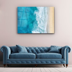May include: A blue and white abstract art print of ocean waves crashing on a sandy beach. The print is framed and hanging on a wall above a blue sofa.