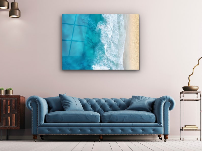 May include: A blue and white abstract art print of ocean waves crashing on a sandy beach. The print is framed and hanging on a wall above a blue sofa.