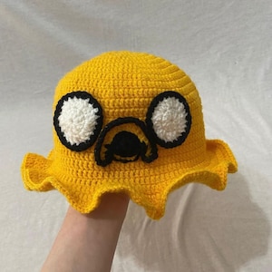 May include: A yellow crocheted bucket hat with a cartoon dog face on the front. The dog has black eyes, a black nose, and a white mouth.