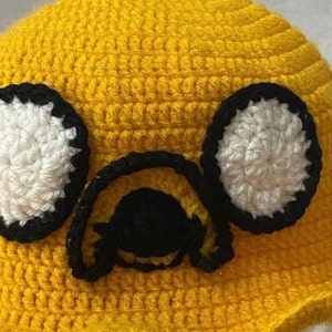 May include: Yellow crocheted hat with black and white crocheted eyes and a black nose. The hat is shaped like a cartoon character.