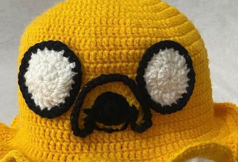 May include: Yellow crocheted hat with black and white crocheted eyes and a black nose. The hat is shaped like a cartoon character.