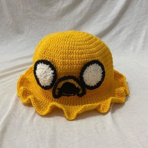 May include: A yellow crocheted bucket hat with a cartoon face on the front. The face has black eyes, a white nose, and a black mouth.