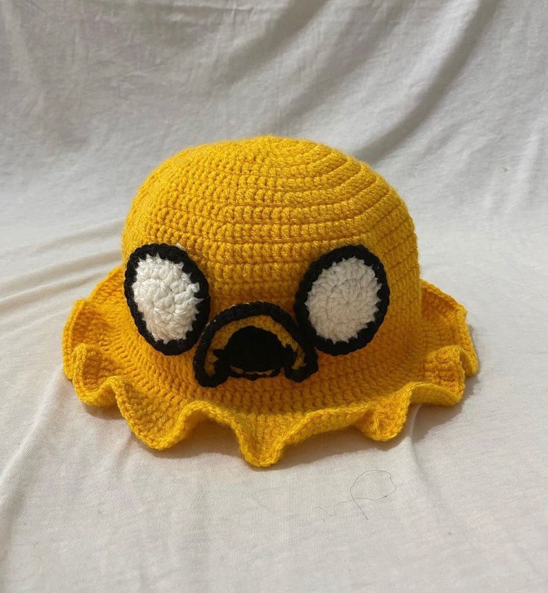 May include: A yellow crocheted bucket hat with a cartoon face on the front. The face has black eyes, a white nose, and a black mouth.