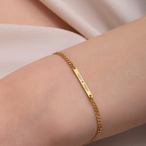 May include: A gold-colored bracelet with a rectangular bar and a delicate chain. The bar has engraved text. The bracelet is worn on a person's arm, against a backdrop of a cream-colored fabric.