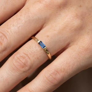 May include: A gold ring with a rectangular, deep blue gemstone. The ring is on a finger, showcasing its delicate design. The band has small decorative cutouts. The ring is a piece of jewelry.