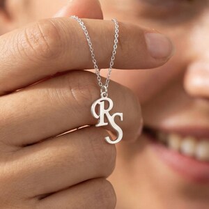 Puede incluir: Un collar plateado con las iniciales "RS" en una fuente cursiva delicada. El colgante cuelga de una fina cadena, creando un accesorio sencillo pero elegante. El collar se muestra sobre un fondo borroso.