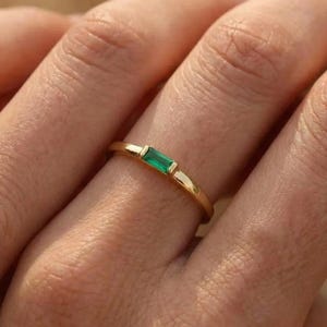 Op de afbeelding: Een gouden ring met een rechthoekige, smaragdgeslepen groene edelsteen. De ring heeft een slanke band. De edelsteen is horizontaal in het midden gezet. Een eenvoudig en elegant ontwerp.