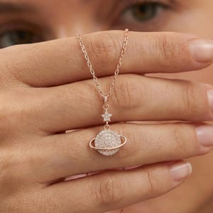 May include: Rose gold necklace featuring a planet pendant with a star accent. The planet is encrusted with small, sparkling stones. The necklace has a delicate chain and a lobster clasp closure. The pendant is approximately 0.75 inches in diameter.