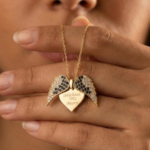 Puede incluir: Collar dorado con un colgante en forma de corazón y alas de ángel. Las alas están adornadas con piedras negras y blancas. El colgante tiene grabados los nombres "Micheal & Mary". El collar es sostenido por una persona.