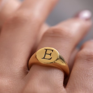 Puede incluir: Un anillo de sello dorado con la letra "E" grabada en negro. El anillo tiene un acabado pulido y una cara clásica de forma ovalada. El anillo se muestra en un dedo.