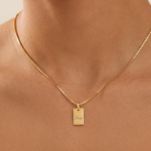 May include: A gold-toned box chain necklace featuring a small, rectangular pendant. The pendant is engraved with the name Isla in a delicate, cursive script. The necklace rests against the neck, showcasing a polished metallic finish and a simple design.