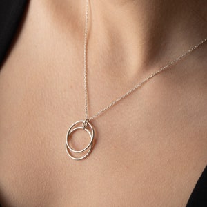 Könnte beinhalten: Eine filigrane Silberkette mit einem Anhänger aus zwei ineinandergreifenden Kreisen. Die feine Kette und der kleine Anhänger schaffen ein minimalistisches Design. Die Halskette wird vor einem dunklen Hintergrund gezeigt, für einen eleganten Look.