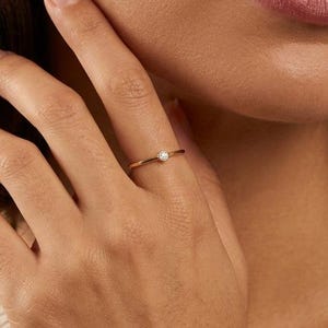 Op de afbeelding: Een dunne gouden ring met een enkele kleine witte parel in het midden. De ring heeft een gladde, gepolijste afwerking en een delicaat, minimalistisch ontwerp dat comfortabel om de vinger zit en een klassieke en verfijnde uitstraling heeft.
