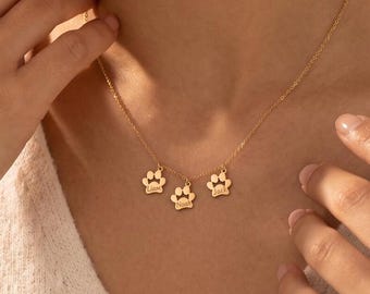 May include: A gold-toned necklace featuring three small, dangling paw print charms. The delicate chain holds the charms in a horizontal arrangement, each shaped like an animal paw with four distinct toe pads and a central pad, creating a simple design.