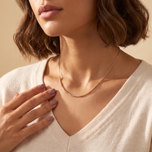 Peut inclure: Un collier à chaîne doré composé d'une série de petites perles métalliques et de courts cylindres espacés régulièrement. Ce collier délicat repose sur le cou, mettant en valeur un motif répétitif de perles et de barres sur la fine chaîne.