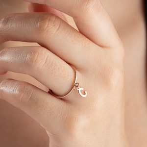 Puede incluir: Un delicado anillo de oro rosa con un pequeño dije colgante. El dije tiene la forma de la letra "C".