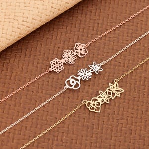 Può includere: Tre delicati braccialetti floreali in oro rosa, argento e oro. Ogni braccialetto presenta una catena con ciondoli a forma di fiore. I braccialetti sono esposti su una superficie marrone strutturata.