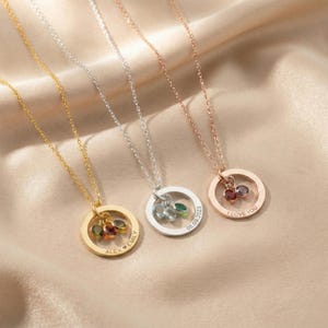 May include: Three necklaces with circular pendants, each featuring gemstones and a fine chain. One is gold-coloured with three gemstones, another is silver with two, and the third is rose gold with two. The pendants have engraved text.