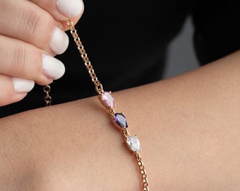 May include: A gold-colored bracelet featuring three teardrop-shaped gemstones in pink, purple, and white. The bracelet is held by a hand with white nail polish, showcasing the delicate chain and gemstone arrangement.