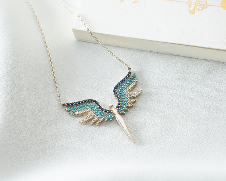 K&ouml;nnte beinhalten: Eine silberne Halskette mit einem blauen und t&uuml;rkisfarbenen Engel-Anh&auml;nger. Der Engel hat Fl&uuml;gel und einen langen, spitzen K&ouml;rper.