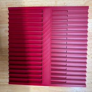 May include: A square, deep red storage tray featuring a series of horizontal, parallel channels or grooves across its surface. The tray is divided into two sections by a central vertical strip with a diagonal, textured pattern.