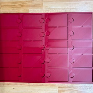 May include: A rectangular dark red plastic organizer tray featuring sixteen individual recessed compartments arranged in a four-by-four grid. Each compartment is rectangular with a small circular cutout on one side for easy access to contents.