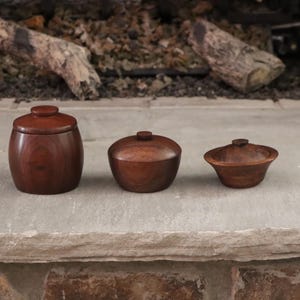 May include: Three dark brown wooden containers with matching lids arranged in a row. The leftmost container is tall and barrel-shaped, the middle one is bowl-shaped, and the rightmost one is a shallow, wide-rimmed bowl with a small lid.