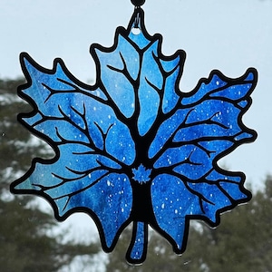 Pode incluir: Um ornamento de folha de bordo de vidro manchado azul e branco com contorno preto. A folha tem um design salpicado e um laço para pendurar na parte superior.