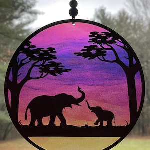 Peut inclure: Un carillon rond en verre coloré avec une silhouette de deux éléphants et des arbres. L'arrière-plan est un dégradé de rose, violet et jaune.