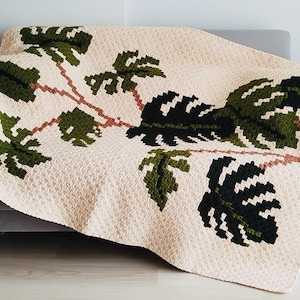 Op de afbeelding: Een gehaakt deken met een witte achtergrond en een groen monstera blad ontwerp. Het deken is over een grijze bank gelegd.