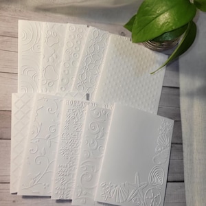 May include: A collection of white embossed cardstock sheets with various patterns, including floral, geometric, and dotted designs. One card features seashells and a starfish. The cards are arranged on a light wood surface.