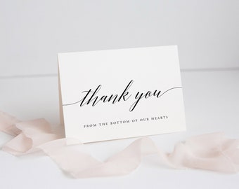 以下が含まれることがあります： 「thank you」と書かれた、エレガントな黒い筆記体のクリーム色のサンキューカード。メインテキストの下には「FROM THE BOTTOM OF OUR HEARTS」と書かれています。淡いピンクのリボンがカードの前にかけられています。