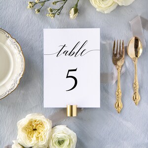 May include: White card with black calligraphy that reads "table 5" on a light blue background with white flowers and gold silverware.