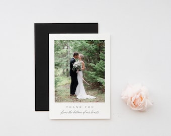 以下が含まれることがあります： 森の中でキスをしているカップルの写真が写った結婚式のサンキューカード。カードには「THANK YOU from the bottom of our hearts」というテキストがエレガントな筆記体で書かれています。黒い封筒とピンクのバラも画像にあります。