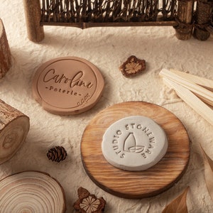 May include: Two round clay stamps, one brown with the text "Caroline - Poterie -" and the other white with the text "Studio Stoneware" and a stylized image of a bottle and bowl.