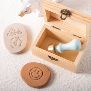 May include: A wooden box with a hinged lid, containing a white ceramic and brass stamp. The box is open and the stamp is visible inside. There are two clay stamps with designs on them, one with the text "Paloma Creations" and the other with a smiley face.