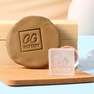 Puede incluir: Un sello de arcilla marrón con el texto "CG POTTERY" presionado en un trozo de arcilla. Un sello transparente con el mismo texto está al lado de la arcilla. El sello está sobre una superficie de madera.