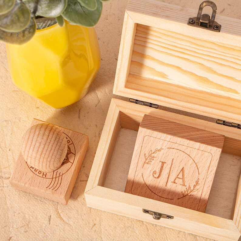 May include: A wooden box with a hinged lid, containing two wooden stamps. One stamp features the initials "J|A" surrounded by a circle and floral designs. The other stamp has the word "Home" and a stylized image of a house.