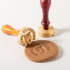 Puede incluir: Dos sellos de latón con mangos de madera. Un sello presenta un diseño de búho y el texto "POTTERY BY CALVIN". El otro sello tiene un borde decorativo. Los sellos se utilizan para crear impresiones en arcilla.