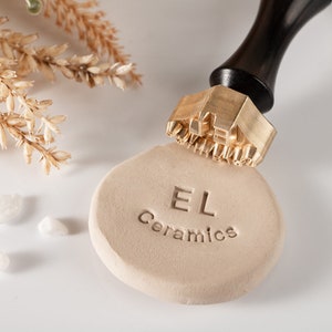 May include: A close-up of a brass stamp with a wooden handle, stamping the words "EL Ceramics" into a beige clay disc.