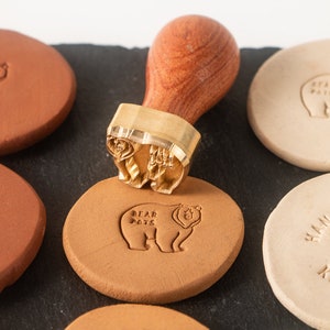 Pode incluir: Um close-up de um carimbo de latão com cabo de madeira com um desenho de urso. O carimbo está sendo usado para criar uma impressão em um disco de argila de cor terracota. O disco tem o texto "BEAR POTS" impresso nele.