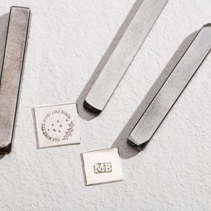 Könnte beinhalten: Vier Metallstempel, zwei lang und zwei kurz, mit unterschiedlichen Designs. Die langen Stempel sind grau und die kurzen Stempel sind silber. Die Designs beinhalten die Worte "Our Scars Like Wings" und die Initialen "MB".