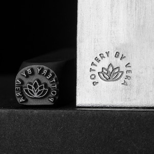 以下が含まれることがあります： 「POTTERY BY VERA」という文字と、スタイリッシュな花柄が刻まれた金属製のスタンプ。このスタンプは、カスタム陶器を作るために使用されます。